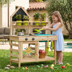 Wooden outdoor play kitchen with girl using scoop, potted plants, and toy vegetables.