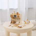 Wooden coffee machine playset with teapot, cups, and snacks on a small table.