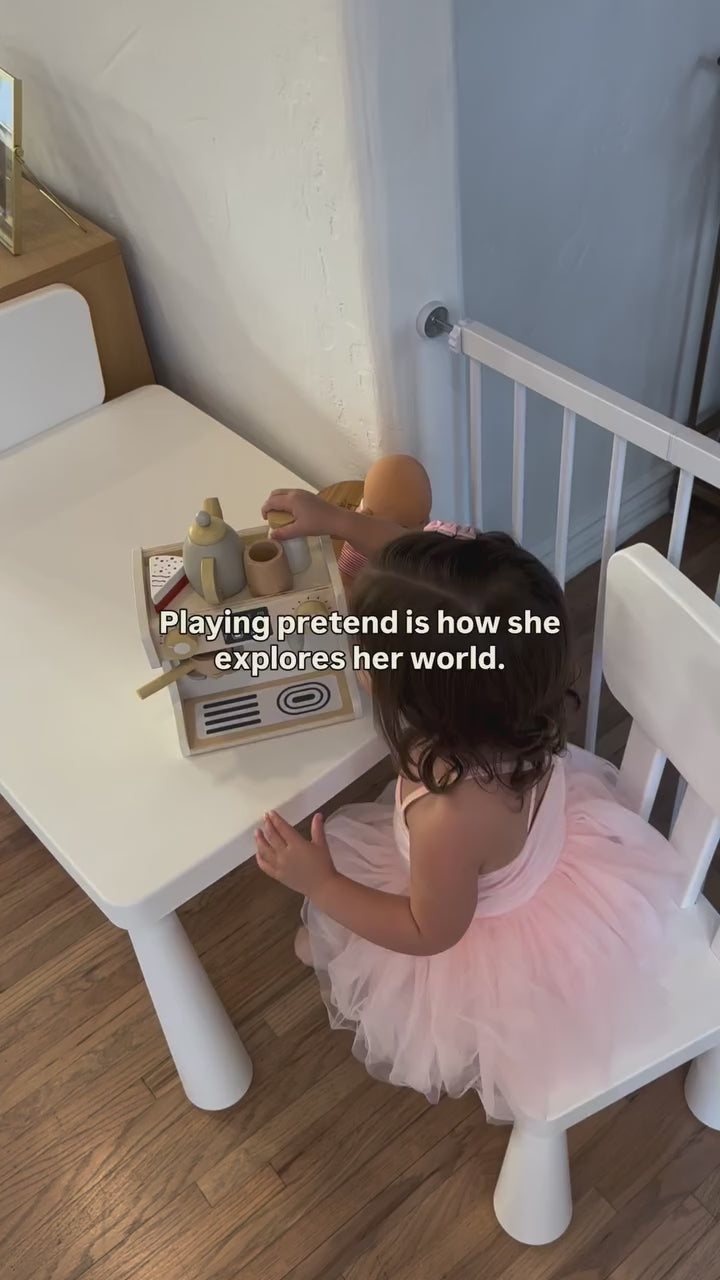 Toddler plays with doll at wooden toy table, exploring world through pretend play.; A toddler plays with a wooden toy kitchen and doll at a small white table.